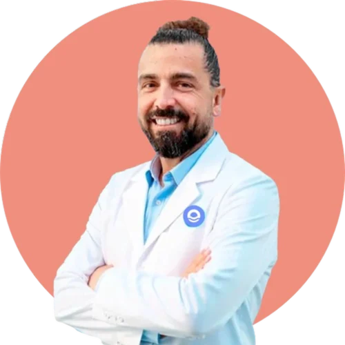 Retrato profesional de busto de un hombre joven con el cabello recogido en un moño alto y barba oscura bien perfilada, que sonríe cálidamente hacia la cámara. Viste una camisa azul claro bajo una bata blanca de profesional sanitario con un logotipo circular azul en la solapa izquierda. Aparece con los brazos cruzados en una postura de confianza y cercanía, recortado sobre un fondo circular de color azul vibrante. Debajo de la imagen, el texto identifica al profesional como Lolo Drago, Director Terapéutico, e incluye un botón azul con el texto "Más información", reflejando el enfoque humano y empático de Bella Mar.