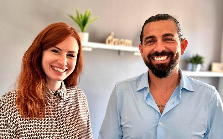 Retrato de primer plano en interior de una mujer joven de cabello pelirrojo con camisa de patrón geométrico junto a Lolo Drago, Director Terapéutico, ambos sonriendo amigablemente frente a una pared gris con estanterías decorativas y la palabra Dream en madera en el centro Bella Mar.