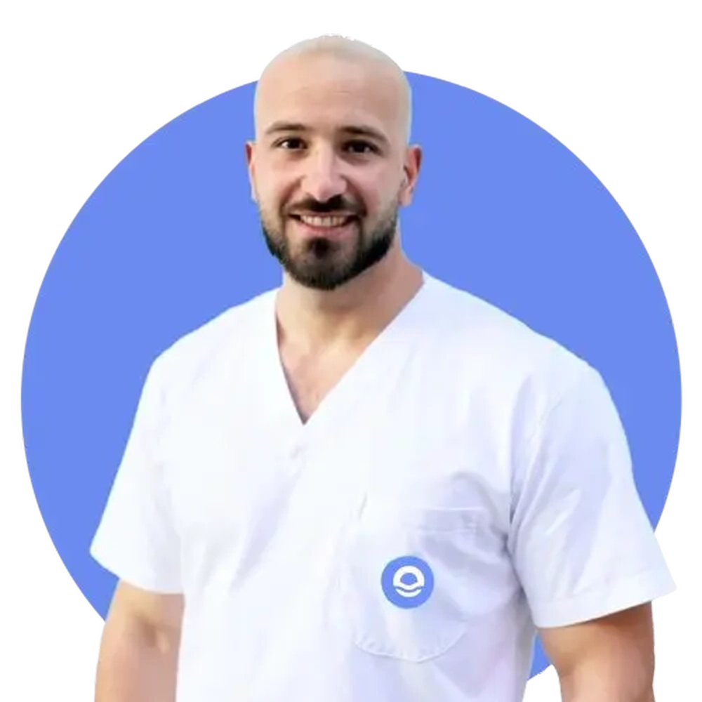 Un retrato profesional de busto de un hombre joven, calvo y con barba oscura bien perfilada, que sonríe amablemente hacia la cámara. Viste un uniforme sanitario de color blanco con cuello en pico, que incluye el logotipo circular azul del centro Bella Mar en el bolsillo del pecho del lado izquierdo. El profesional aparece recortado sobre un fondo circular de color azul vibrante que resalta su figura y transmite una imagen de confianza y estabilidad. Bajo la imagen, el texto identifica al profesional como Marc Vidiella, Monitor, destacando su rol esencial en el soporte diario de los residentes.