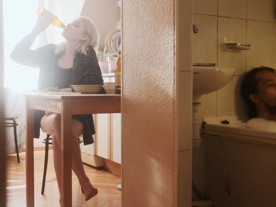 Una imagen con un fuerte contraste. A la izquierda, se observa a una mujer bebiendo directamente de la botella, dando pequeñas señales de un posible alcoholismo, a su derecha, la que entendemos que es su pareja necesitando fumar continuamente para poder paliar con la situación siendo el, por consecuencia de esto, otro drogodependiente
