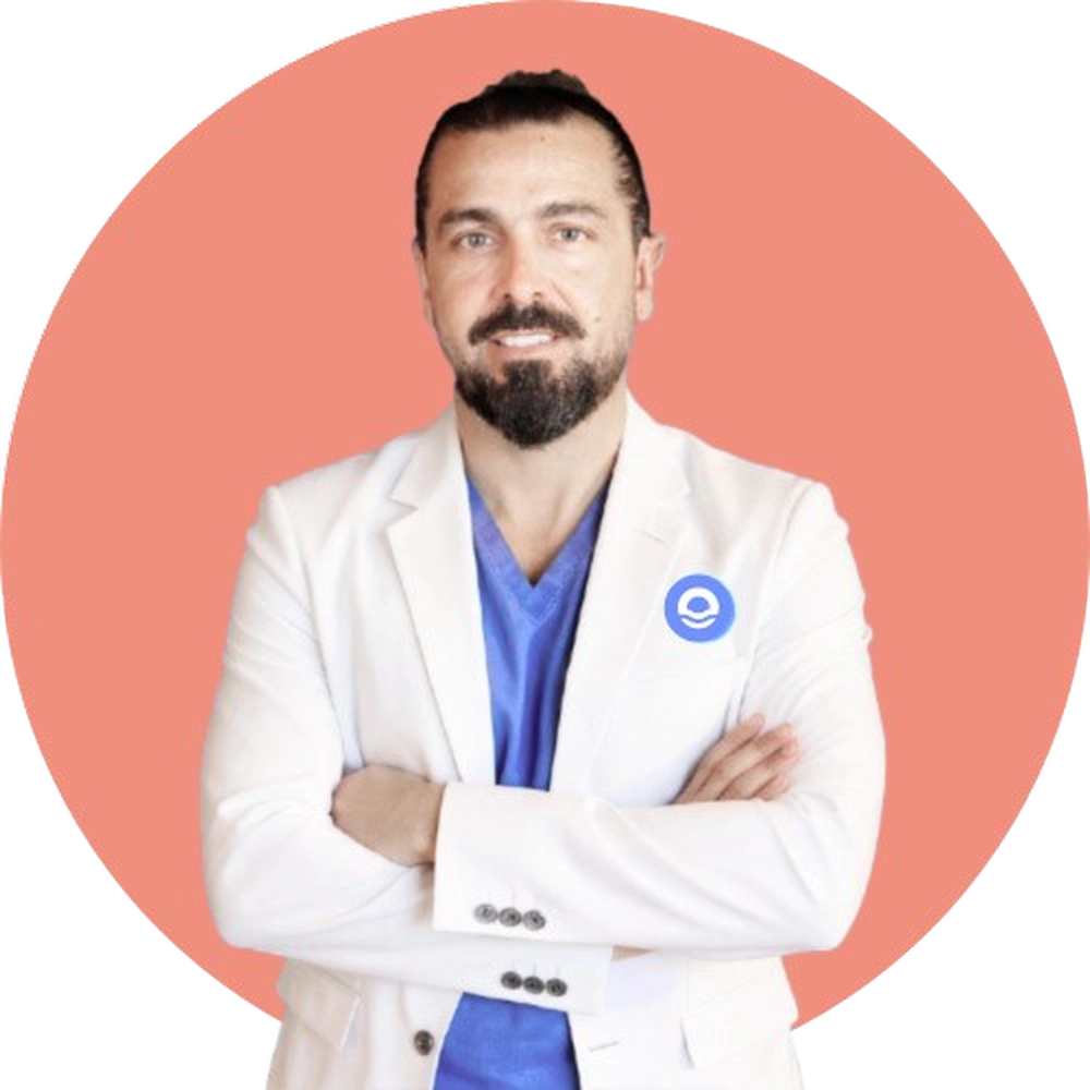 Retrato profesional de busto de un hombre joven con el cabello recogido en un moño alto y barba oscura bien perfilada, que sonríe cálidamente hacia la cámara. Viste una camisa azul claro bajo una bata blanca de profesional sanitario con un logotipo circular azul en la solapa izquierda. Aparece con los brazos cruzados en una postura de confianza y cercanía, recortado sobre un fondo circular de color azul vibrante. Debajo de la imagen, el texto identifica al profesional como Lolo Drago, Director Terapéutico, e incluye un botón azul con el texto "Más información", reflejando el enfoque humano y empático de Bella Mar.