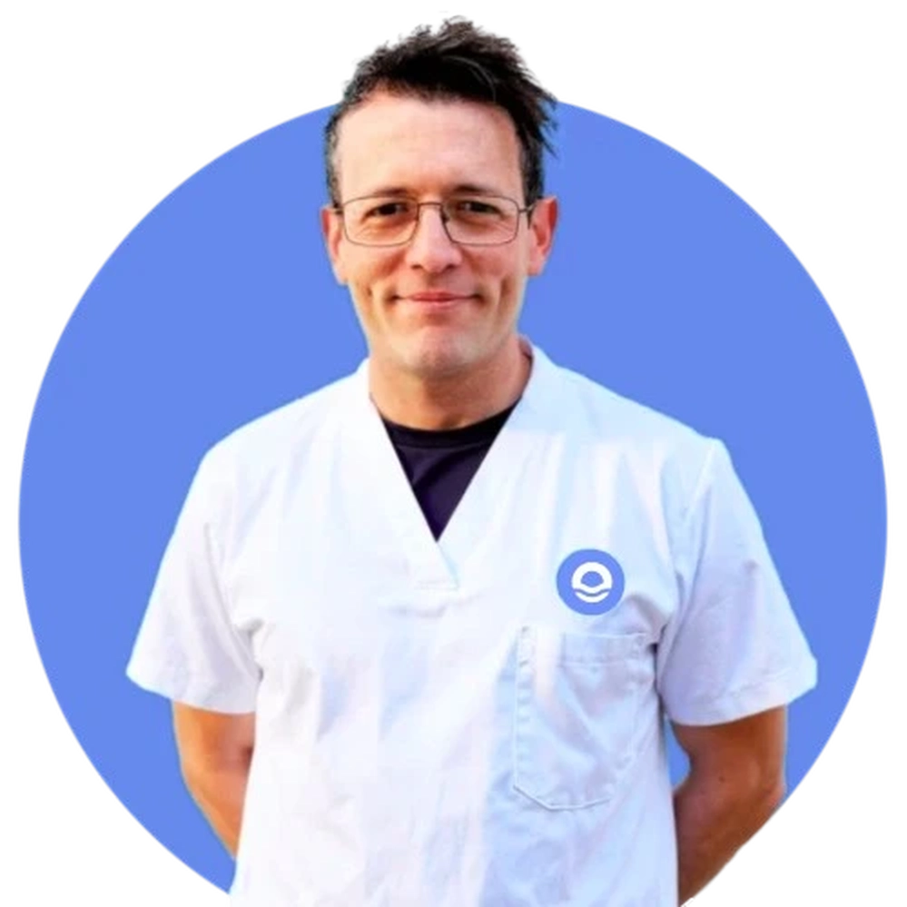 Retrato profesional de busto de un hombre adulto con gafas de montura fina, cabello oscuro corto y expresión amable con una leve sonrisa. Viste un uniforme sanitario blanco de cuello en "V" que muestra una camiseta oscura debajo. En el pecho, a la altura del corazón, resalta el logotipo circular azul característico de Bella Mar. La imagen está contenida dentro de un círculo azul vibrante sobre fondo blanco. Bajo el retrato, el texto identifica al profesional como Enrique Sanz Martínez, destacando sus cargos de Sociosanitario, Técnico Superior en Integración Social y Coordinador de Admisiones.