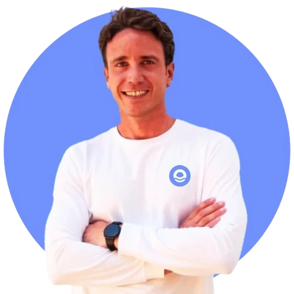 Un retrato profesional de busto de un hombre joven llamado Daniel Jover, con el cabello castaño corto y una sonrisa abierta y acogedora. Viste una camiseta técnica de manga larga en color blanco, que luce el logotipo circular azul de Bella Mar en la parte superior izquierda del pecho. Daniel posa con los brazos cruzados, mostrando un reloj deportivo negro en su muñeca izquierda. La imagen está enmarcada dentro de un círculo de color azul vibrante sobre un fondo blanco limpio, manteniendo la coherencia estética de las presentaciones del equipo del centro.