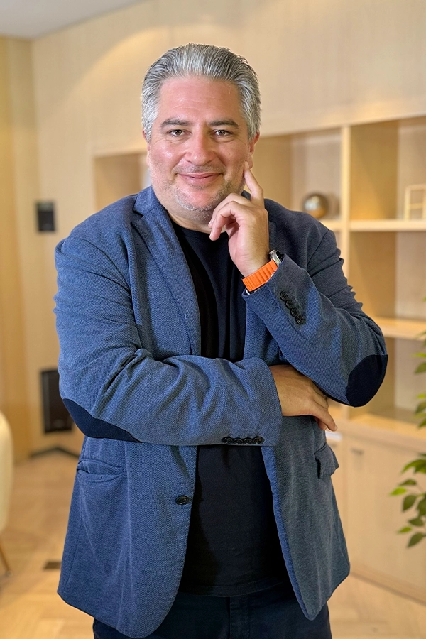 Un retrato profesional de plano medio del Dr. Méndez, un hombre de mediana edad con cabello canoso y barba corta y cuidada. Viste una chaqueta tipo blazer de color azul jaspeado sobre una camiseta negra, transmitiendo una imagen de profesionalidad relajada. Posa con los brazos cruzados, apoyando su mano derecha en la mejilla en una actitud reflexiva y amable. En su muñeca destaca un reloj con una llamativa correa de color naranja vibrante. El fondo muestra una estantería de madera clara con objetos decorativos, sugiriendo un despacho moderno, cálido y acogedor dentro de las instalaciones de Bella Mar.