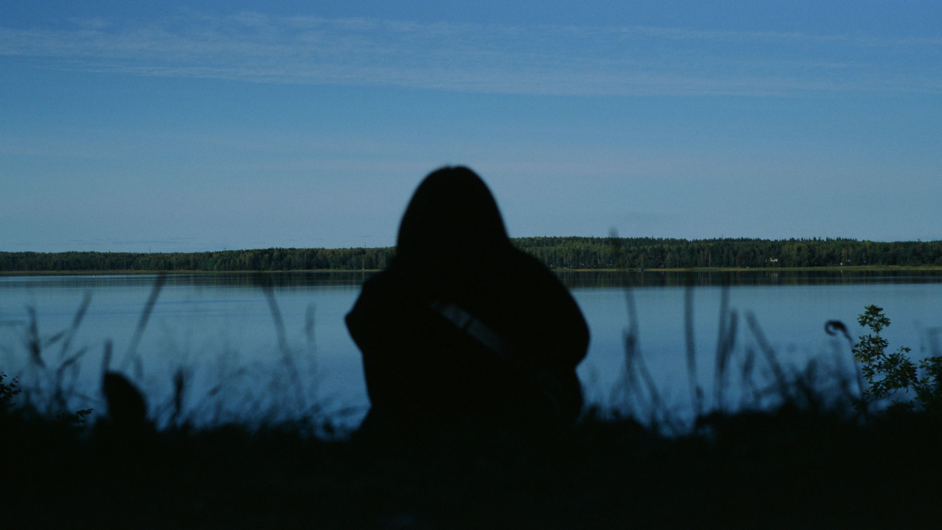 Una fotografía capturada en condiciones de baja luminosidad y a contraluz, donde se observa la silueta oscura de una persona sentada de espaldas, con la cabeza ligeramente inclinada hacia adelante. La figura se encuentra en la orilla de un cuerpo de agua sereno, posiblemente un lago o un río ancho, bajo un cielo de color azul profundo y crepuscular. En el horizonte se distingue una franja continua de bosque oscuro. En el primer plano, algunas briznas de hierba silvestre aparecen desenfocadas, acentuando la sensación de introspección, soledad y la carga emocional que conllevan los daños a largo plazo de las adicciones tratados en Bella Mar.