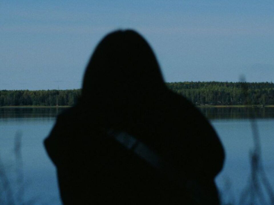 Una fotografía capturada en condiciones de baja luminosidad y a contraluz, donde se observa la silueta oscura de una persona sentada de espaldas, con la cabeza ligeramente inclinada hacia adelante. La figura se encuentra en la orilla de un cuerpo de agua sereno, posiblemente un lago o un río ancho, bajo un cielo de color azul profundo y crepuscular. En el horizonte se distingue una franja continua de bosque oscuro. En el primer plano, algunas briznas de hierba silvestre aparecen desenfocadas, acentuando la sensación de introspección, soledad y la carga emocional que conllevan los daños a largo plazo de las adicciones tratados en Bella Mar.