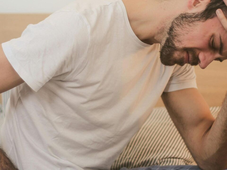 Una fotografía de plano medio que muestra a un hombre joven sentado en el borde de una cama desordenada en una habitación con iluminación tenue. Viste un pijama compuesto por una camiseta blanca de manga corta y pantalones de cuadros azules. El hombre aparece encorvado, con su mano derecha presionando su frente en un gesto de intenso dolor, frustración o desesperación, mientras su mano izquierda descansa sobre su espalda baja. La expresión de su rostro, parcialmente oculta, refleja agotamiento físico y mental. Al fondo se aprecia el cabecero de madera de la cama y una mesita de noche blanca, creando una atmósfera de soledad y reflexión íntima.