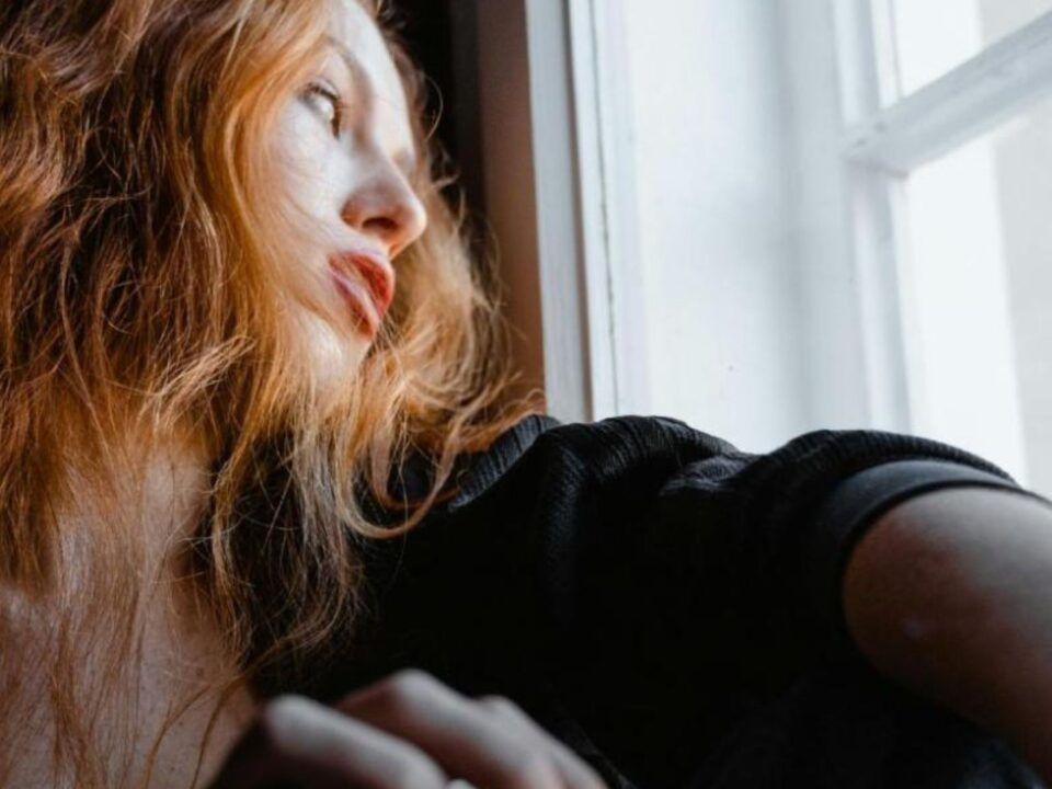 Una fotografía de plano medio capturada desde un ángulo bajo que muestra a una mujer joven de cabello pelirrojo largo y ondulado. Se encuentra en una habitación con poca luz, situada junto a una ventana por la que entra una claridad fría y difusa. La mujer viste una prenda negra y mira hacia el exterior con una expresión de profunda melancolía, ausencia y cansancio. En primer plano, su mano derecha aparece ligeramente desenfocada cerca del marco de la ventana, subrayando la sensación de encierro y la desconexión con el mundo real que caracteriza a las adicciones digitales tratadas en Bella Mar.