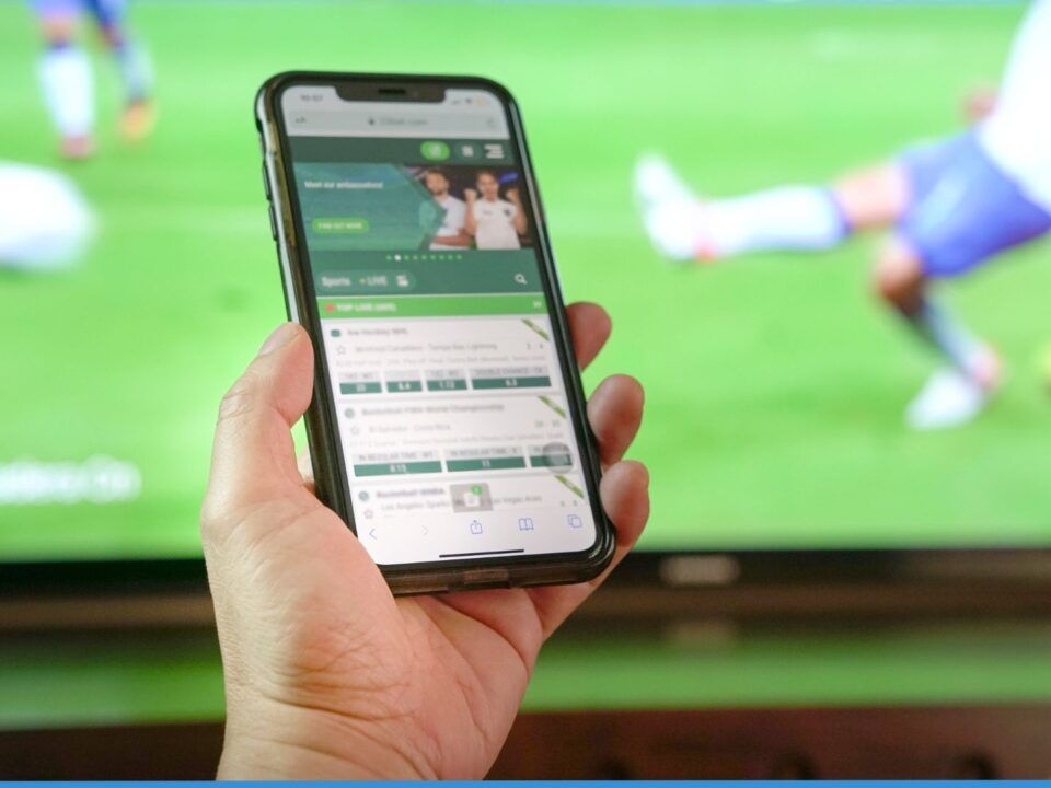 Una fotografía de primer plano que muestra la mano de una persona sosteniendo un teléfono móvil inteligente (smartphone) frente a una pantalla de televisión de grandes dimensiones. En la pantalla del teléfono se visualiza una aplicación de apuestas deportivas con listas de cuotas y mercados de eventos en vivo. Al fondo, la televisión muestra un partido de fútbol desenfocado donde se ve a un jugador en acción sobre el césped verde. La imagen captura la conexión inmediata entre el consumo de contenido deportivo y la facilidad de acceso al juego digital, ideal para ilustrar los disparadores de la ludopatía en Bella Mar.