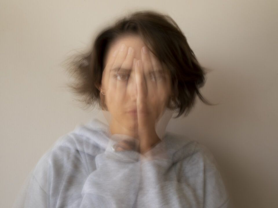 Retrato de plano medio con un efecto de larga exposición o doble exposición que crea un desenfoque de movimiento. En la imagen, una persona aparece con las manos frente al rostro, pero la distorsión visual hace que las facciones y las manos se multipliquen en capas semitransparentes. Proyectando una sensación de inestabilidad, agitación interna y la dificultad de mantener el enfoque en medio de una crisis de ansiedad.