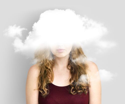 Una composición conceptual que muestra el torso y rostro de una mujer vestida con una prenda color granate. El elemento central es una nube blanca y densa que flota directamente sobre la parte superior de su cara, ocultando sus ojos y frente. Representación literal del término "niebla mental" o bloqueo emocional. Los mechones de cabello ondulado caen a los lados, mientras que los labios permanecen visibles, sugiriendo una persona que está presente físicamente pero desconectada de la realidad. 